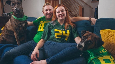psychology alum mara sargent and her partner dressed in university of oregon gear sit on a couch with their dogs